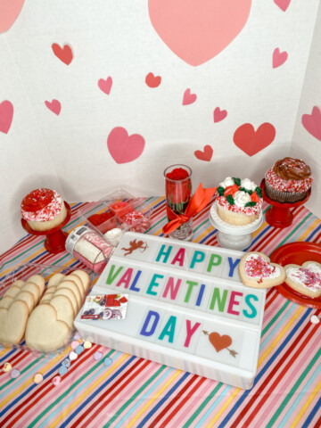 valentine's day sign with colorful letters, unfrosted heart shaped cookies, decorated cupcakes on a striped tablecloth
